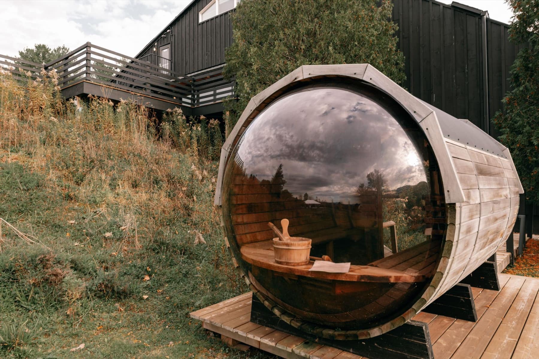 A cedar barrel sauna with a round glass end sits on a small deck beside tall dry grass.