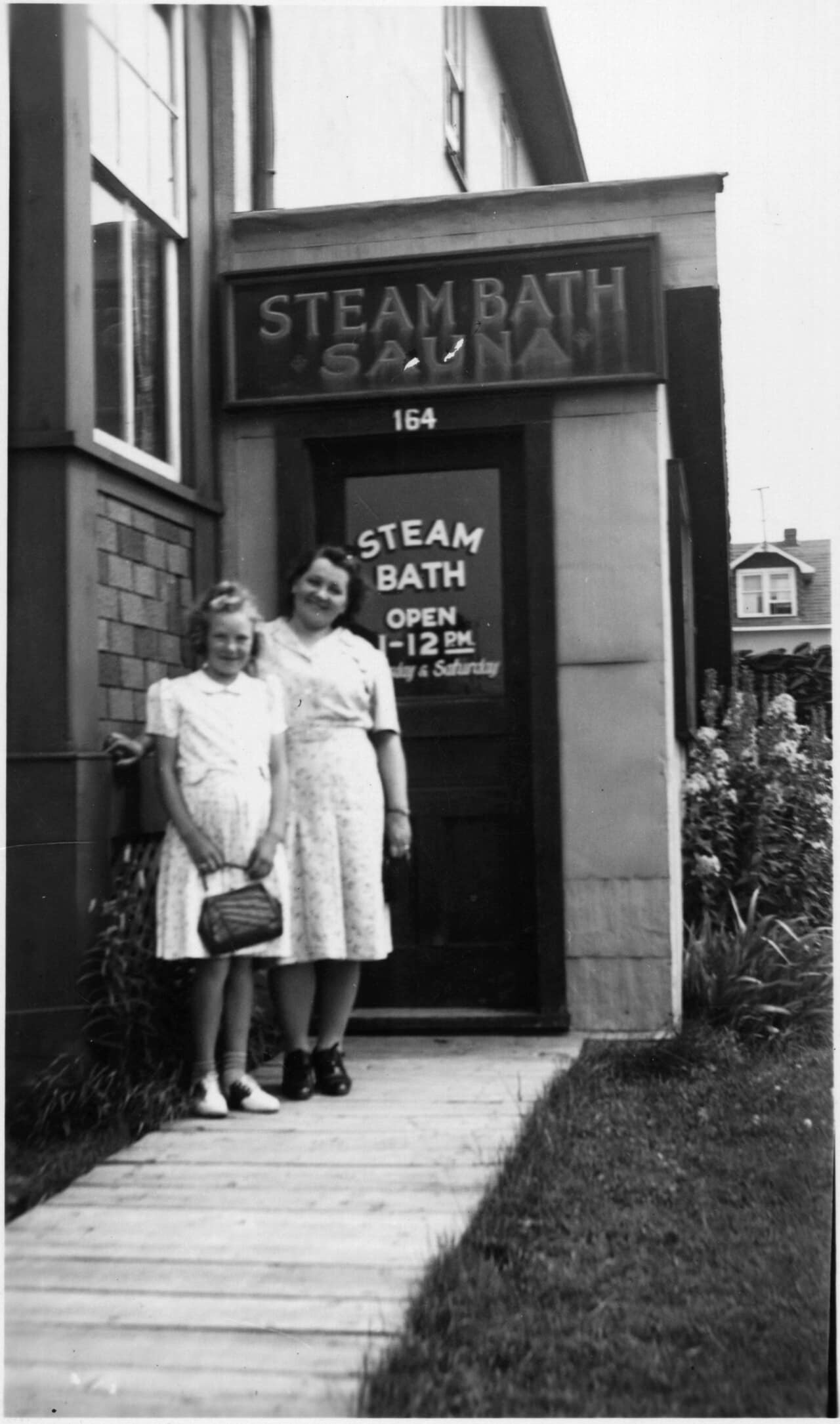 Bertta Kaja and Helvi Syrja stand at the door of a public steam bath in Cochrane, Ontario, in 1946, under a sign reading Steam Bath Sauna.
